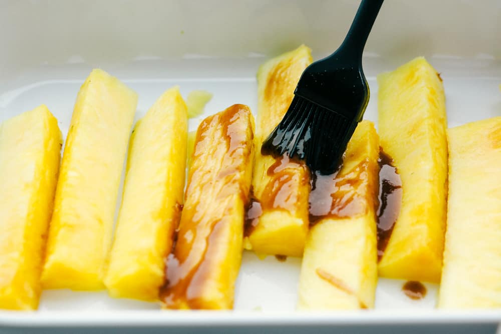 Pineapple spears lined up in a white dish getting brushed with a caramelized glaze. 