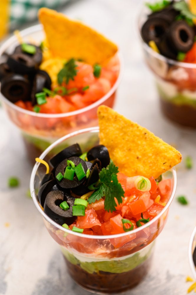 Individual cups with seven layer dip inside with black olives, tomatoes, scallions and a chip on top. 