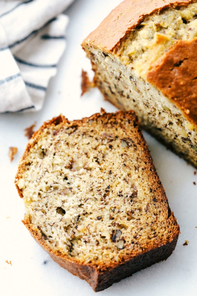 Una barra de pan de plátano con una rebanada de pan de plátano tendido sobre la encimera blanca. 