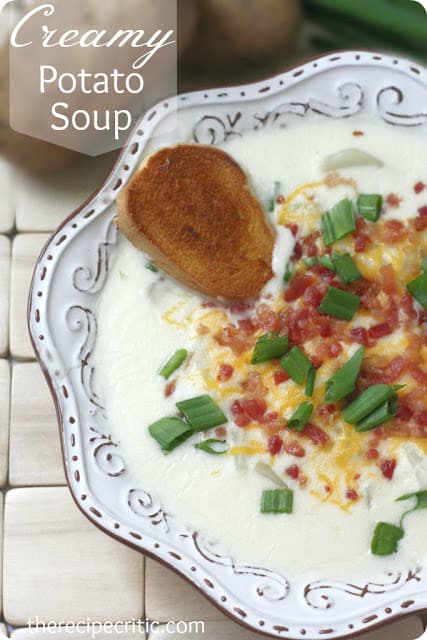 Creamy potato soup in a white dish garnished with a bread, shredded cheese and scallions. 