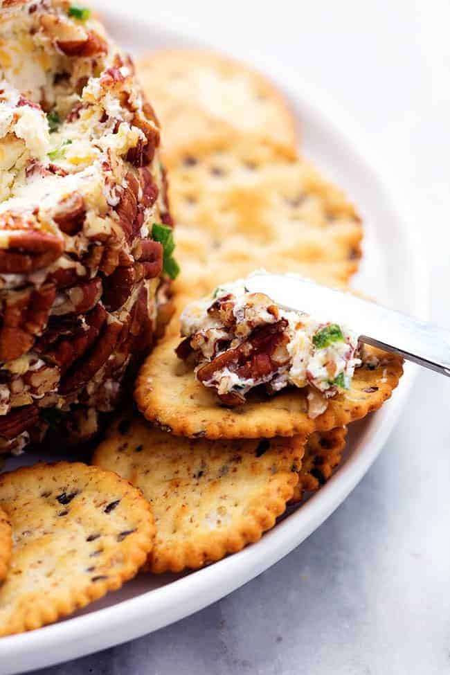 Bacon ranch cheeseball on a white plate with crackers being spread with a cheeseball knife.