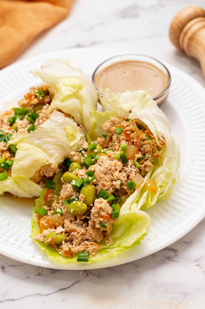 Two lettuce wraps on a white plate with a glass bowl of dipping sauce. A pepper grinder lays beside the plate.