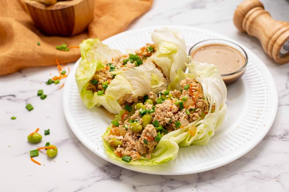 Two lettuce wraps on a white plate with a glass bowl of dipping sauce. A pepper grinder lays beside the plate on the right.