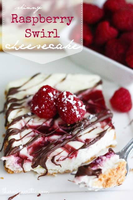 Raspberry Swirl cheesecake square with a chocolate drizzle, 2 raspberries on top and a sprinkle of powdered sugar on a white plate. A small square white bowl of raspberries is beside the cheesecake. A fork with a bite of cheesecake lays beside the piece.