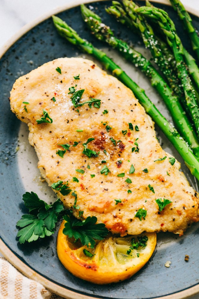 Baked Parmesan garlic chicken with asparagus on a plate garnished with a lemon and parsley. 