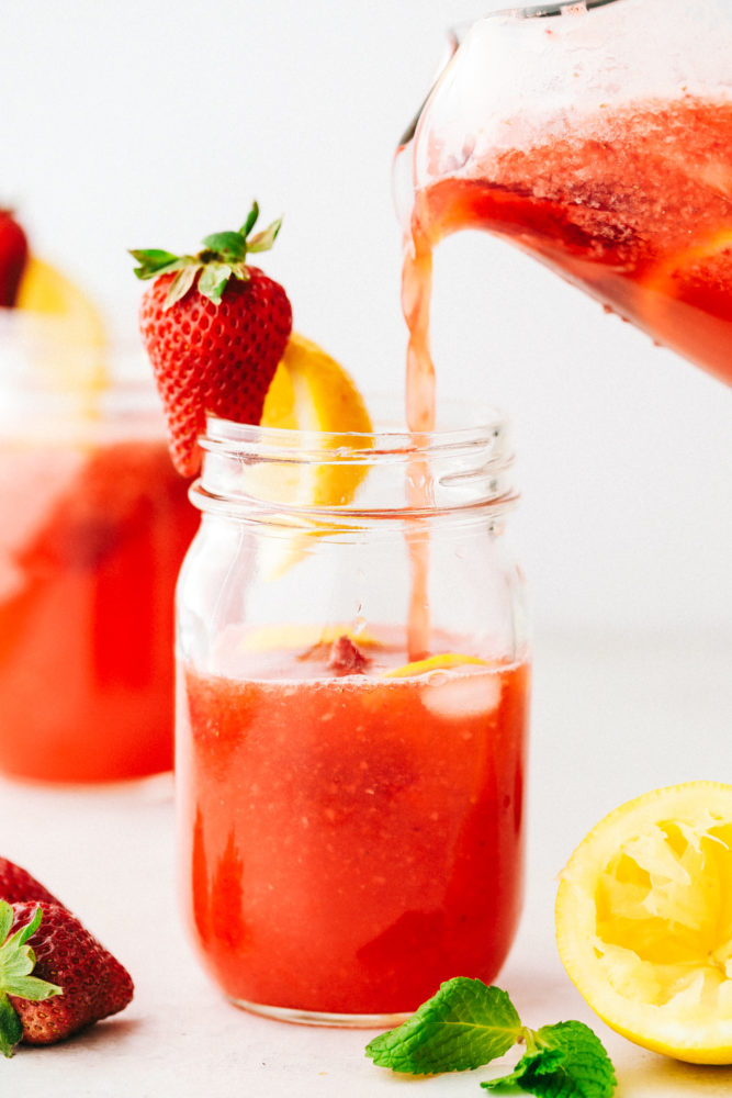A mason jar with strawberry lemonade being poured into it and a strawberry with a lemon stuck to the side. 