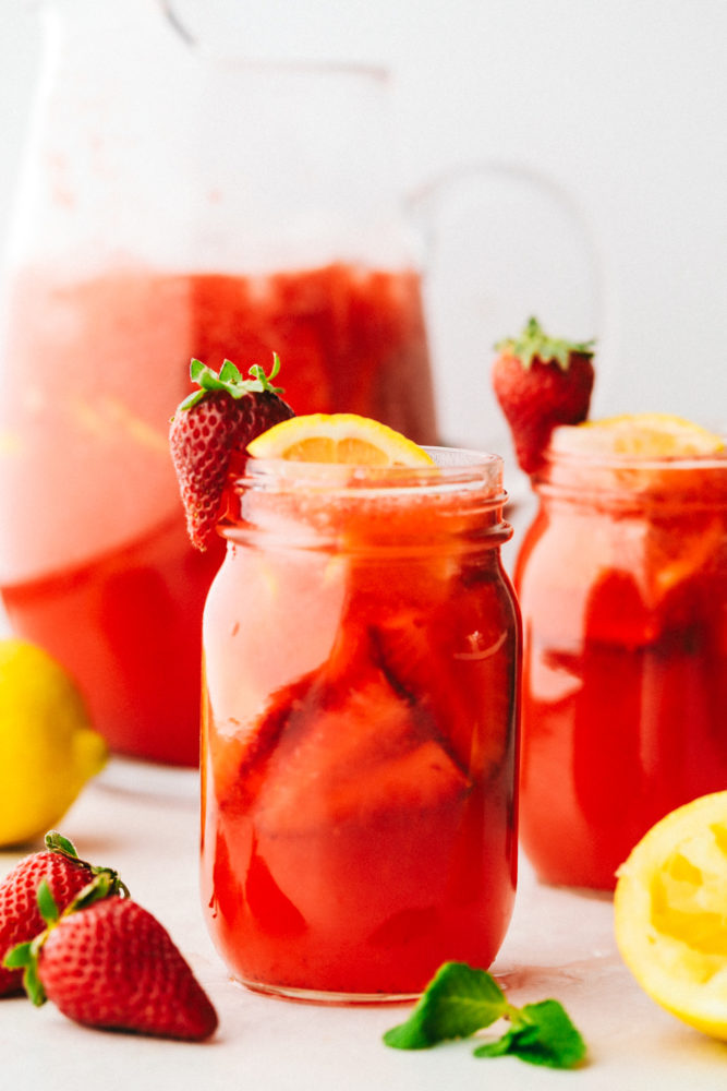 Homemade strawberry lemonade ina a mason jar garnished with lemons and strawberries with a pitcher of lemonade in the background faded out and another mason jar with strawberry lemonade. 