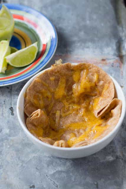 a white bowl with a tortilla in it covered in melted cheese. A colorful plate with lime slices sits beside the bowl on the gray tray.