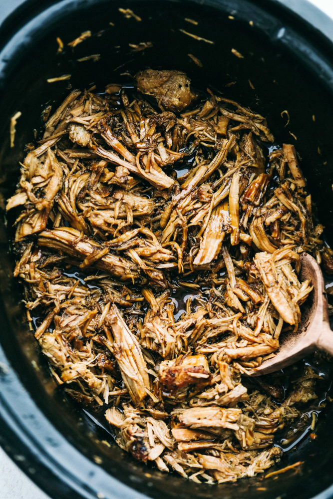 Shredded pork in a slow cooker being stirred with a wooden spoon. 