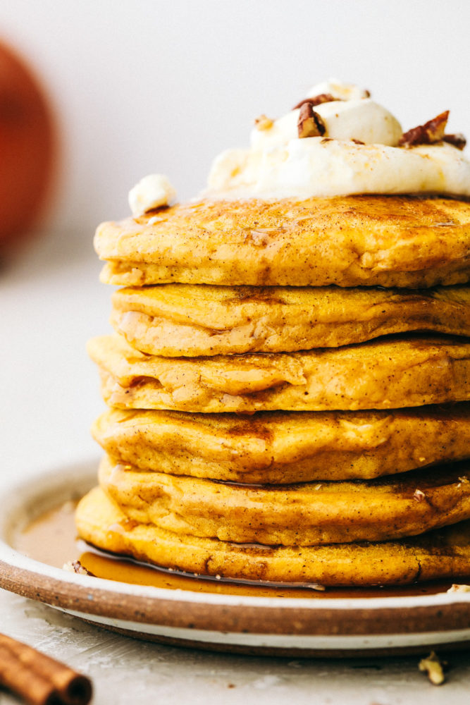 Close side view of a stack of pumpkin spice pancakes. Whipped cream and pecans are garnished on the top pancake.