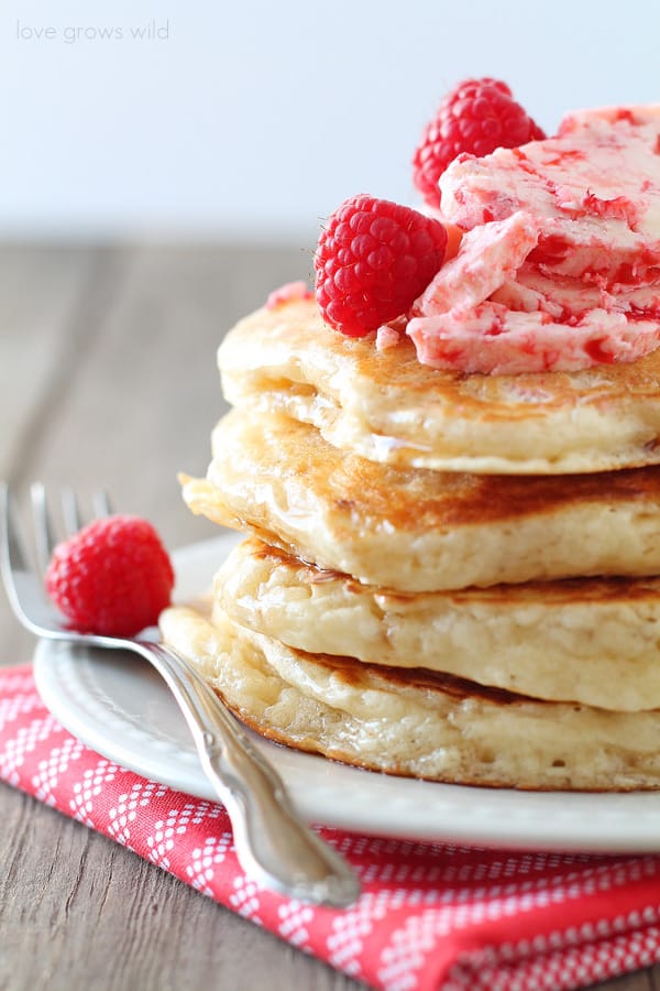 Close up of a stack of pancakes with Raspberry Butter on top with a metal fork on the side, all placed on a white plate. 