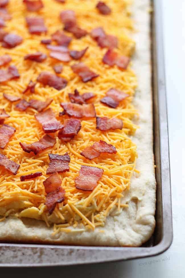 Fresh grated cheese and sliced bacon that has been placed over the scrambled egg on the pizza crust. 
