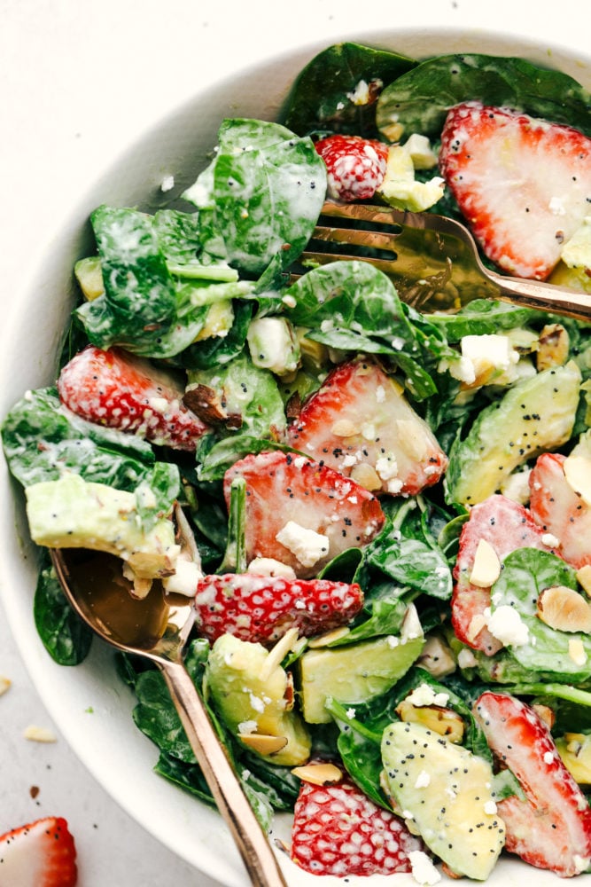 Ensalada de espinacas de aguacate y fresa con aderezo de semillas de amapola mezcladas y colocadas en un tazón con un tenedor y una cuchara. 