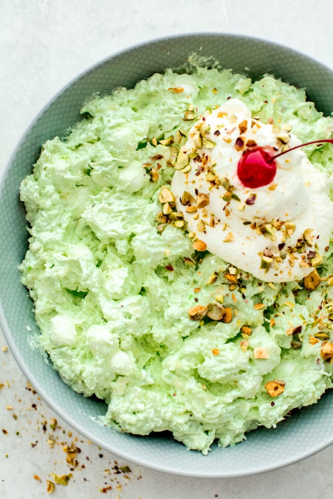 Pistachio pudding salad in a bowl topped with whipping cream and a cherry on top. 