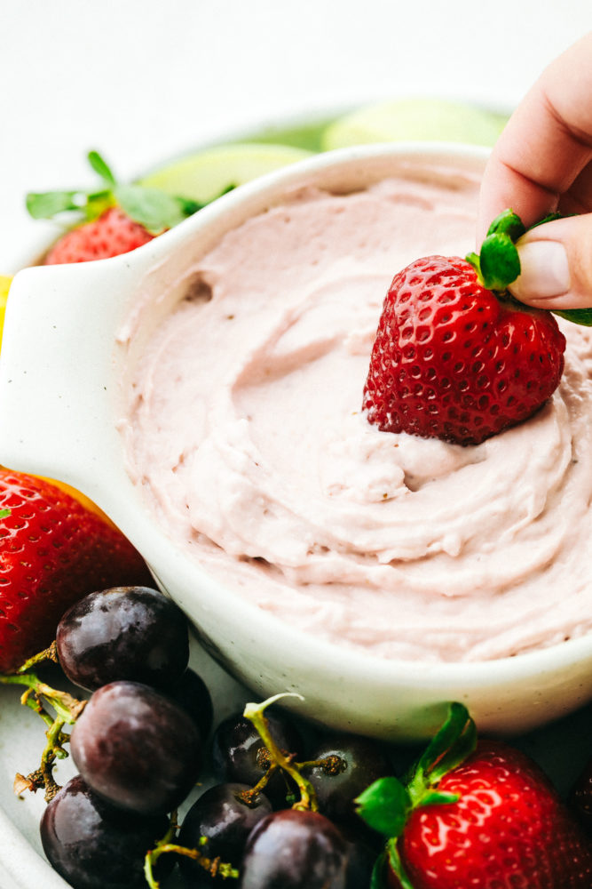 A hand dipping a large strawberry in a white dish of Strawberry Cream Cheese Fruit Dip. 