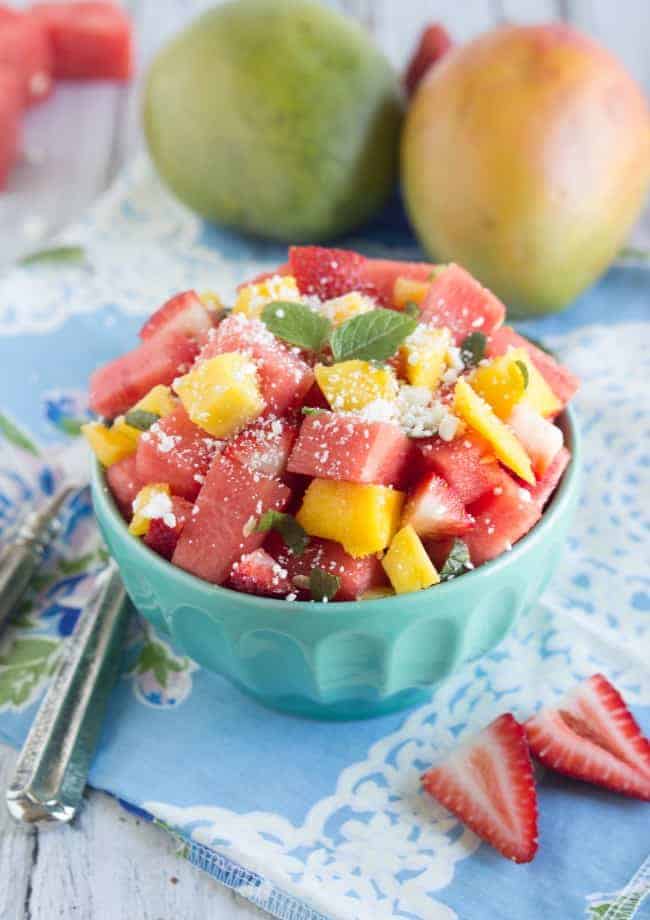 Fresh watermelon and feta salad in a light blue bowl with whole mangos in the background. 