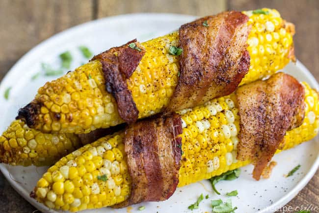 Grilled bacon corn on the cob laying on a white plate.