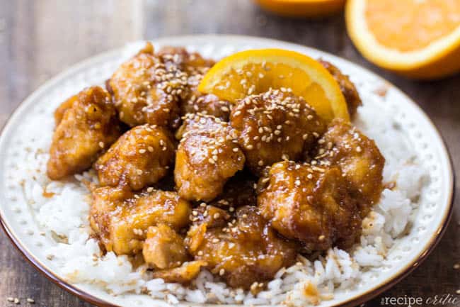 Baked orange chicken on a plate of white rice with an orange slice on the side. 
