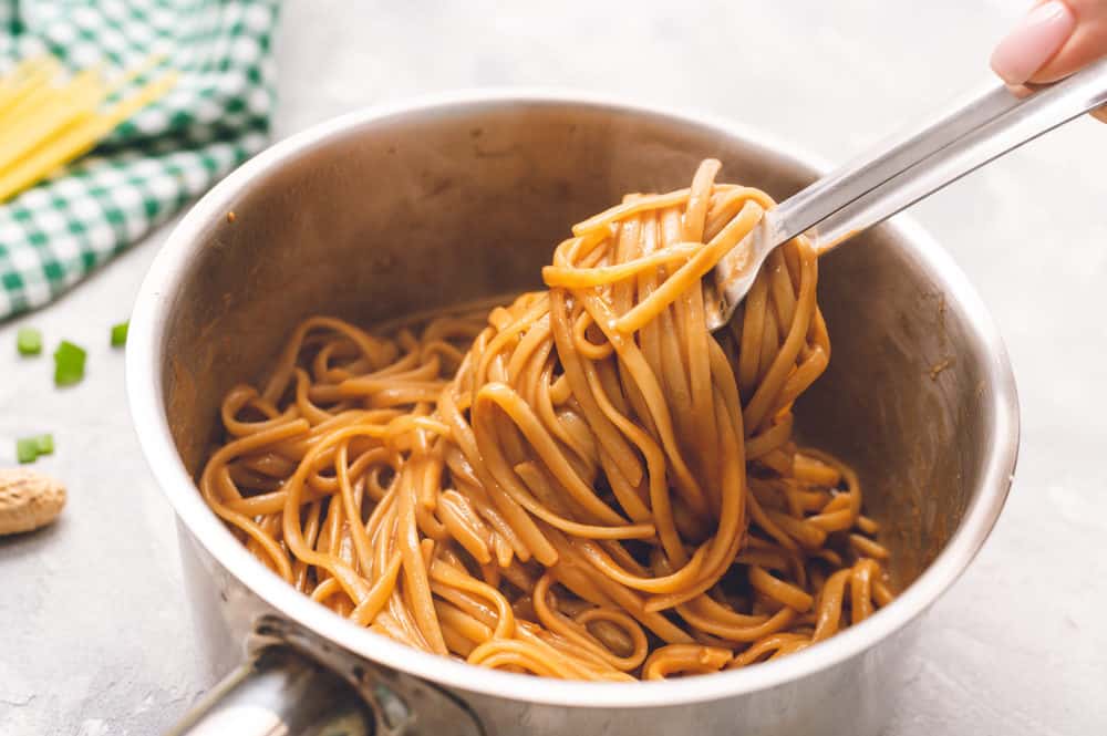 Thai peanut sesame noodles cooked in a pan with tongs grabbing it out of the pan. 