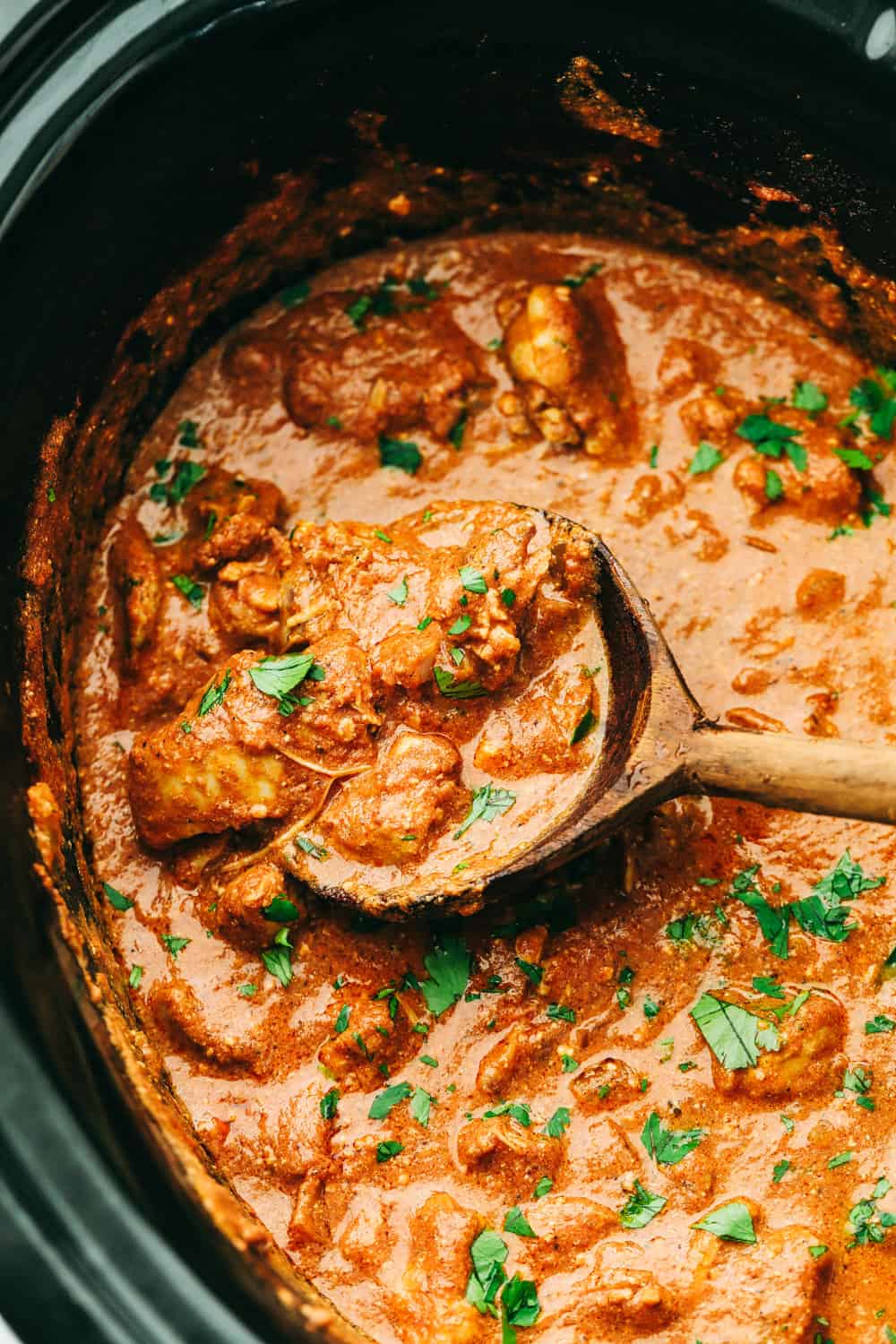 Chicken tikka masala in a slow cooker with a wooden spoon scooping up chicken in the sauce.