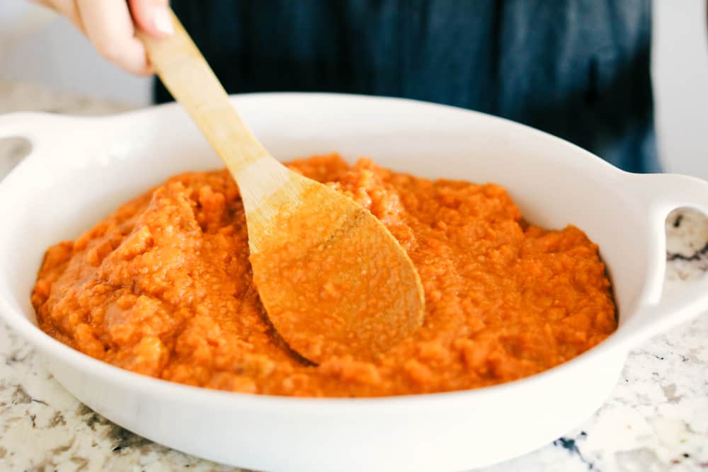 sweet potatoes being smoothed and layered into a casserole dish with a wooden spoon