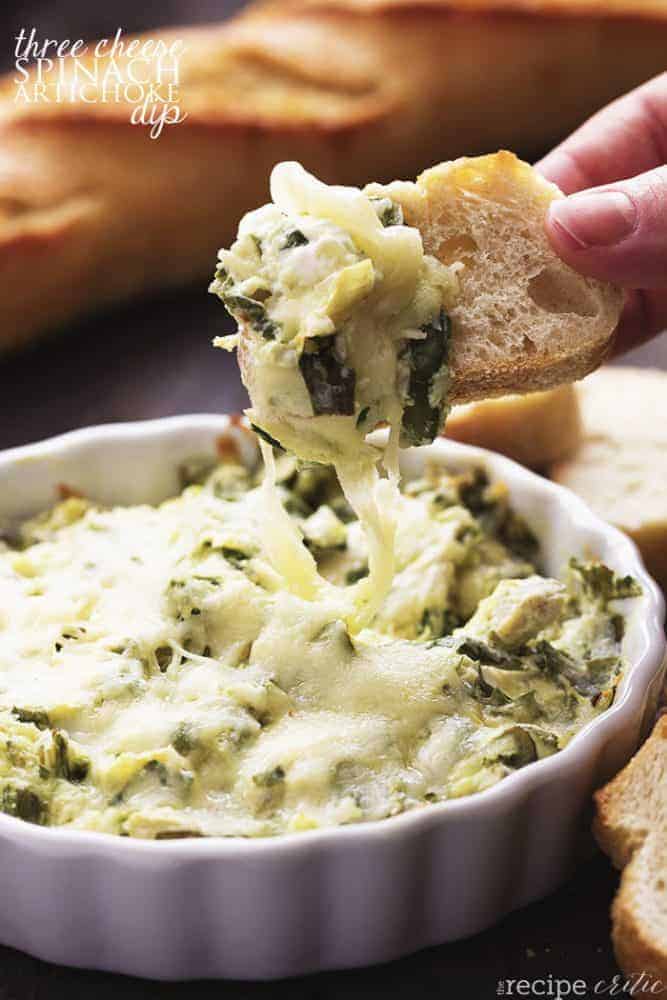 Three cheese spinach artichoke dip in a white bowl with a piece of bread dipping into the bowl.