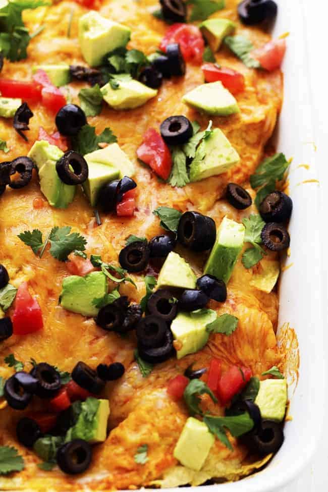 stacked enchilada in a white cooking dish with avocado and black olives toppings.