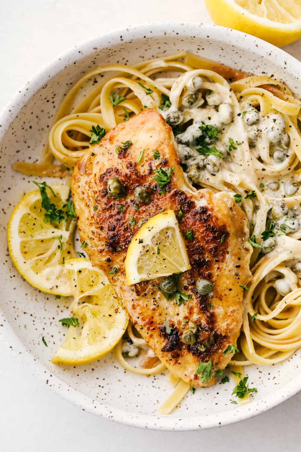 Chicken breasts with lemon piccata sauce on a bed of noodles in a white bowl. 