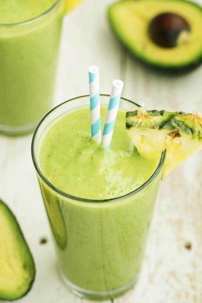 Avocolada smoothie in a glass with pineapple garnish and blue and white striped straws.