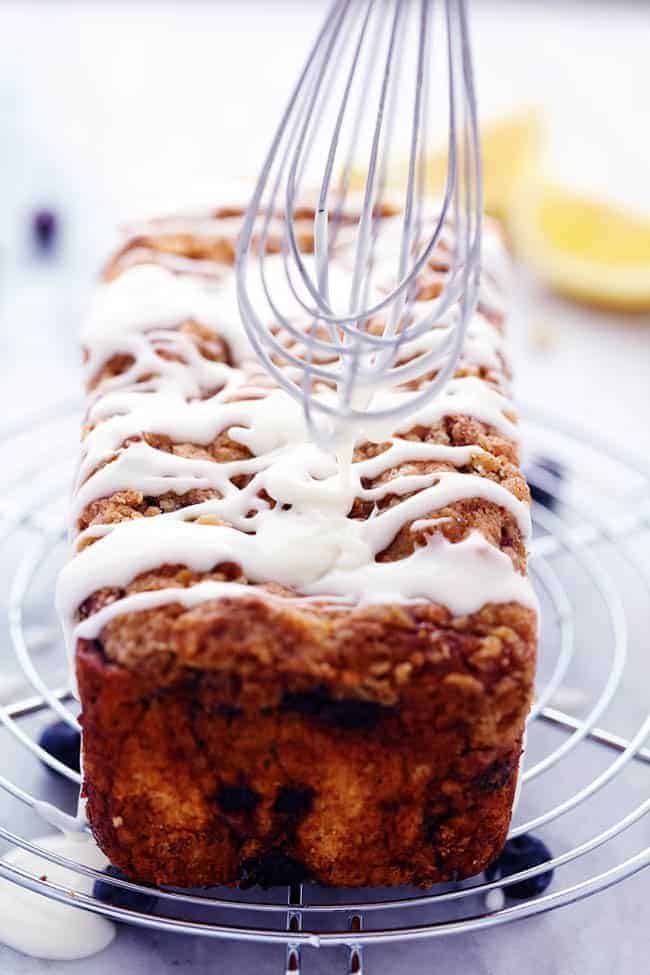 Blueberry pie bread on a cooling rack with lemon glaze being spread by a whisk.