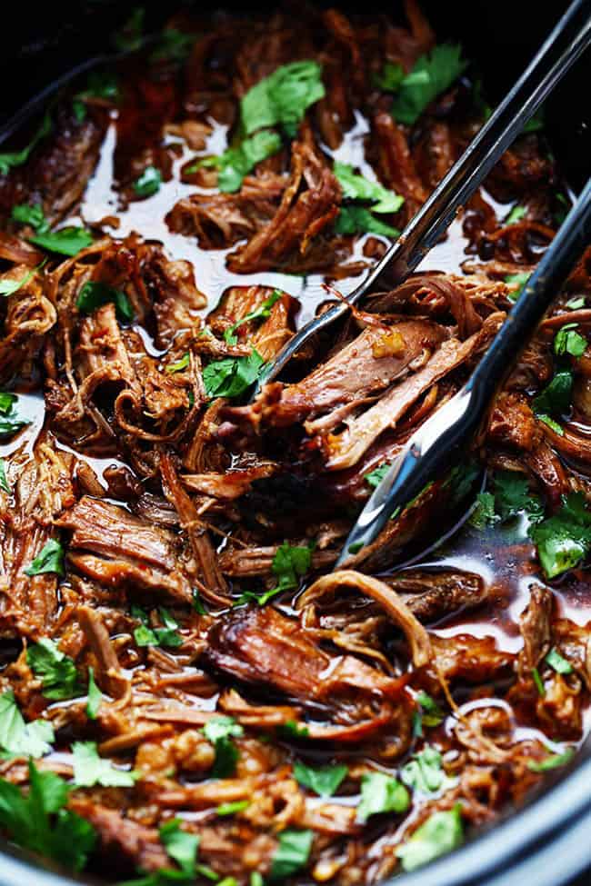 Barbacoa de ternera en una olla de cocción lenta con pinzas tirando un poco de la carne que se muestra en la foto.