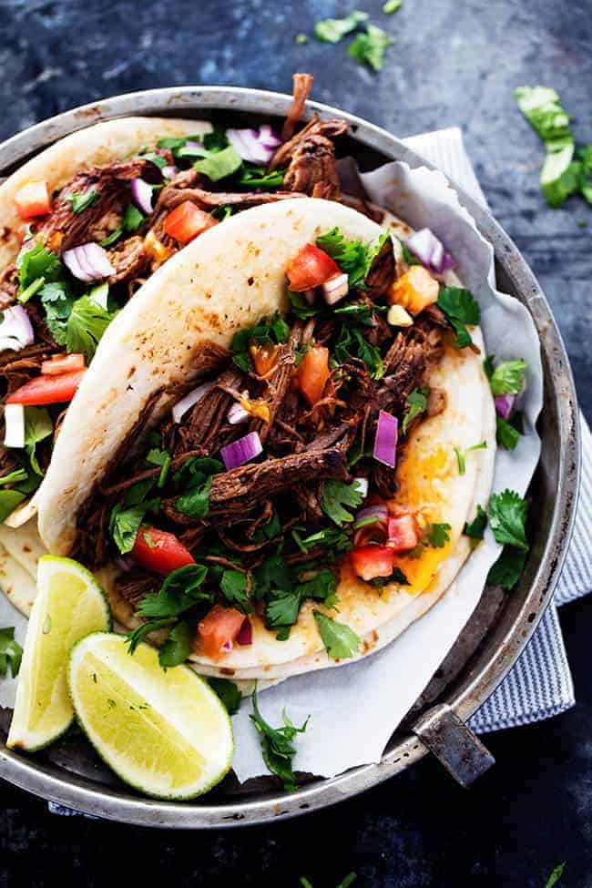 Barbacoa en un taco con tomates, cebollas y lechuga encima con limas a un lado. 