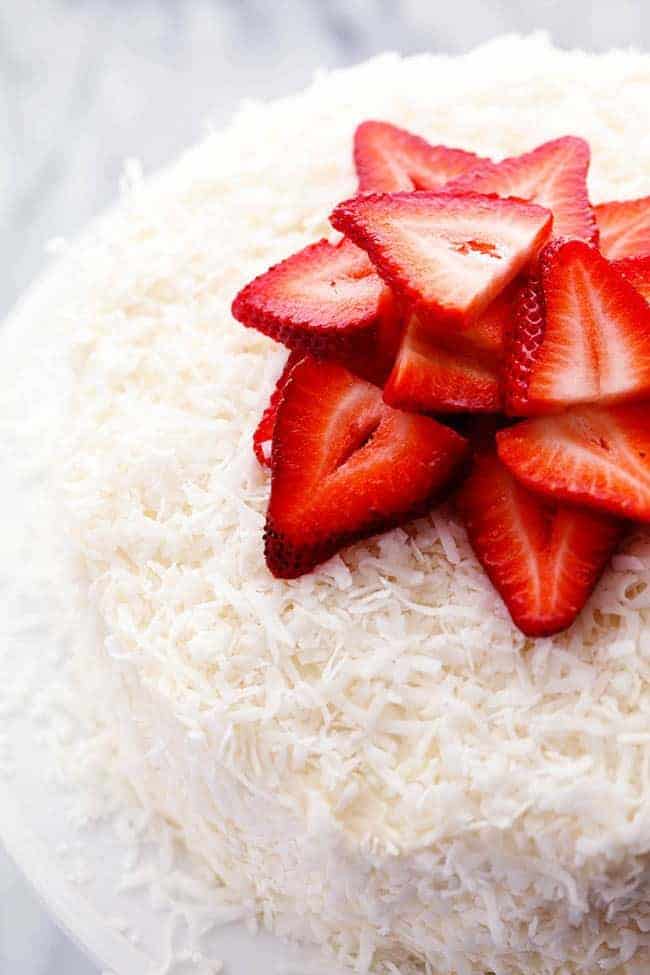 Erdbeer-Kokos-Sahne-Torte mit Kokos-Frischkäse-Zuckerguss und frisch geschnittenen Erdbeeren auf einem weißen Tortenständer. 