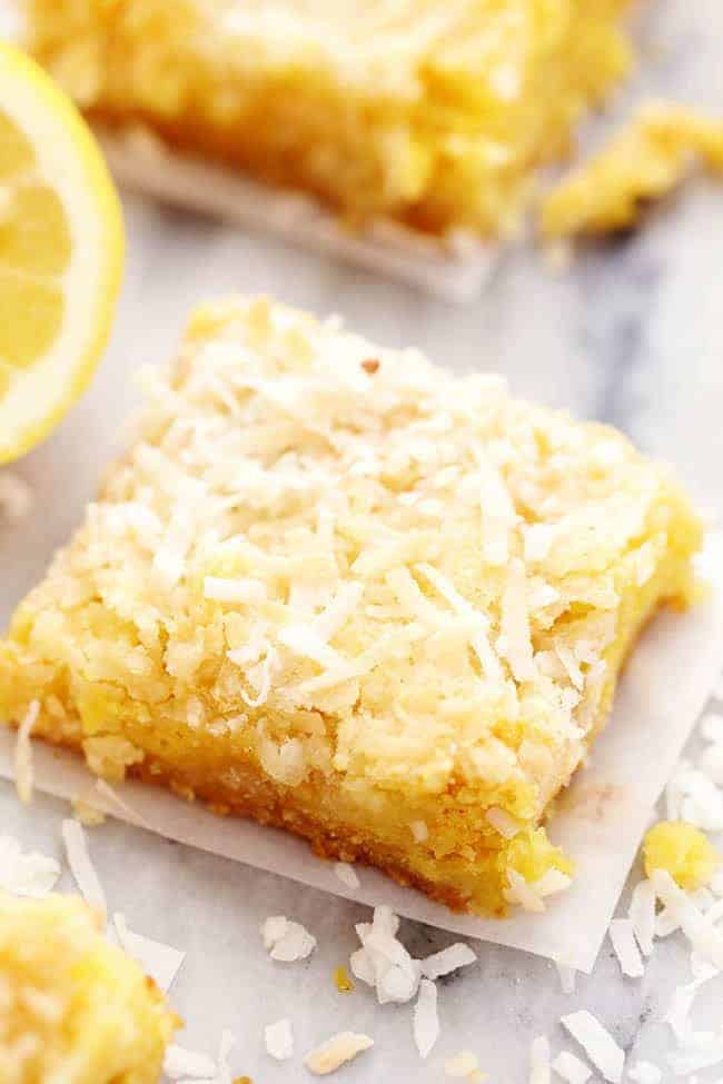 One lemon coconut butter bar on parchment paper with a fresh cut lemon in the background.