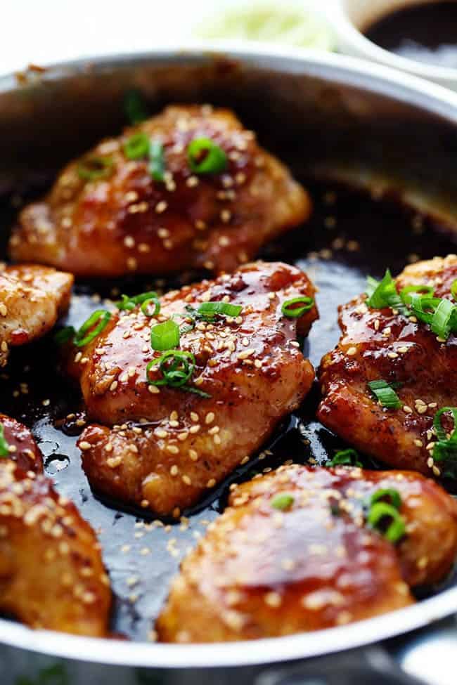 Sticky asian glazed chicken cooking in a skillet with scallions and sesame seed garnished on top.