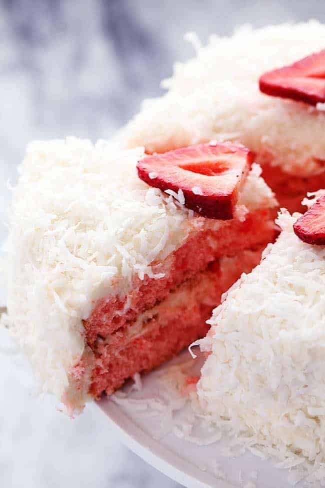 Strawberry Coconut Cream Cake with Coconut Cream Cheese Frosting on a white cake stand with a slice being removed. 