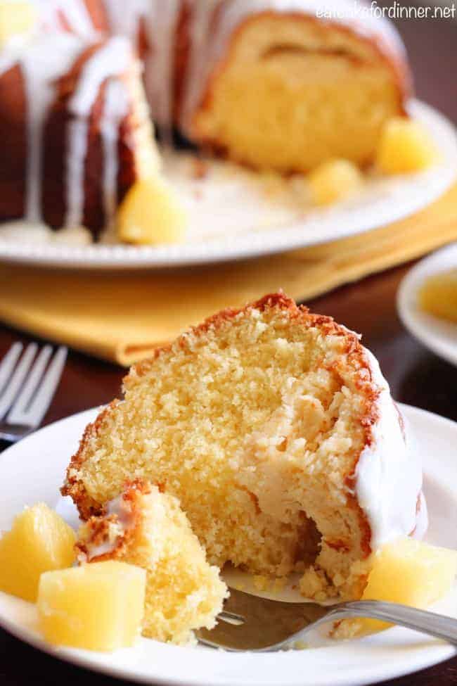 A slice of Pineapple Cheesecake Cake on a white plate with a metal fork. The rest of the cake is on a white platter in the background.  