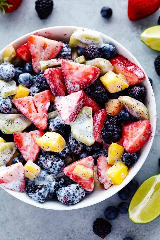 Creamy poppyseed fruit salad in a white bowl with fresh lemons, strawberries, blueberries, and blackberries on the side