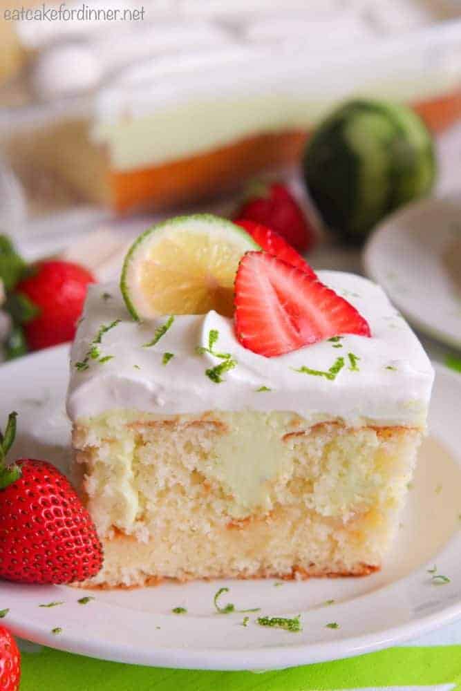 Key Lime Poke Cake on a white plate with fresh strawberries and lime on top. 