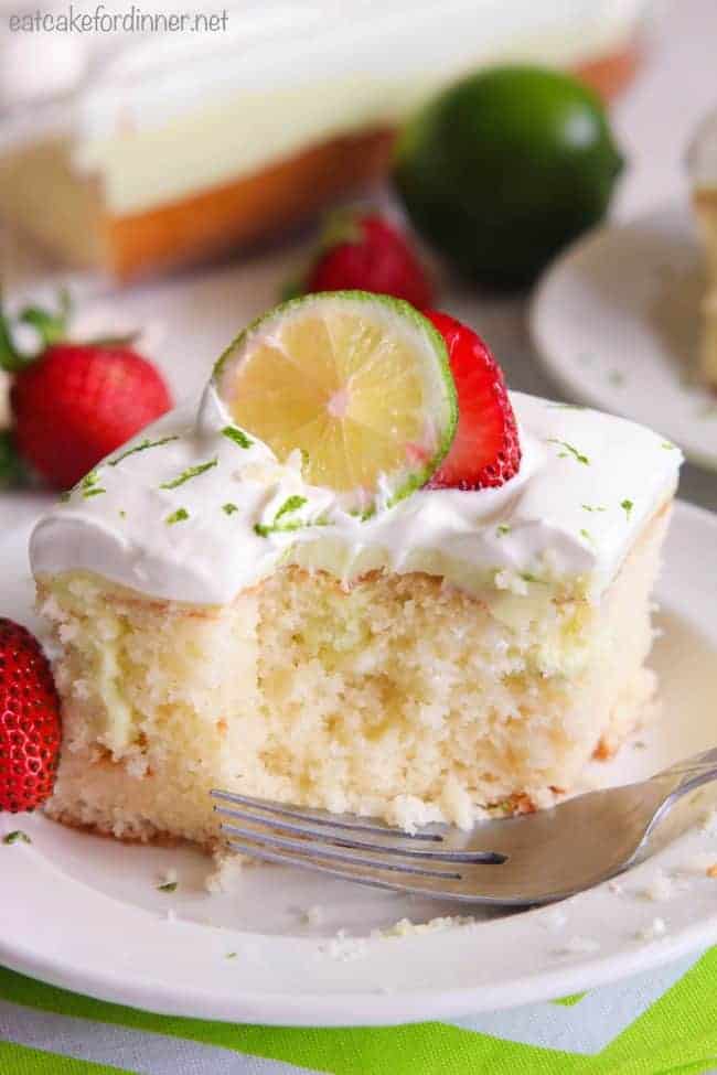 Key Lime Poke Cake on a white plate with fresh strawberries and lime on top. 