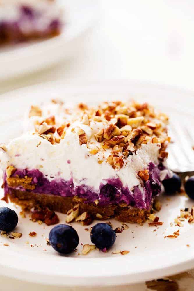 No Bake Creamy Blueberry Pecan Square auf einem weißen Teller mit einer Metallgabel und frischen Blaubeeren als Beilage. 