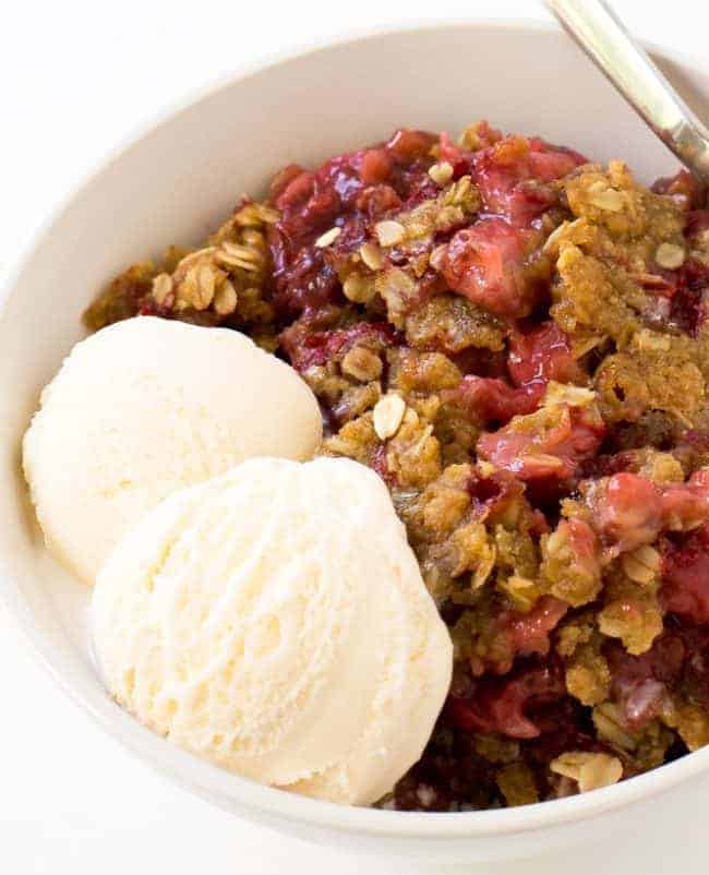 Strawberry Crisp in a white bowl with two scoops of ice cream and a metal spoon. 