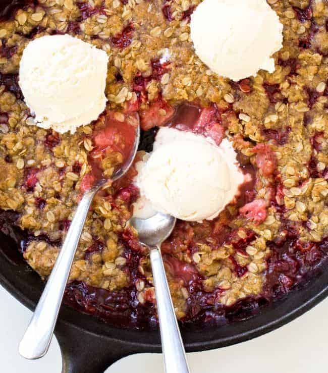 Strawberry Crisp in a large black skillet with three scoops of ice cream and two metal spoons. 