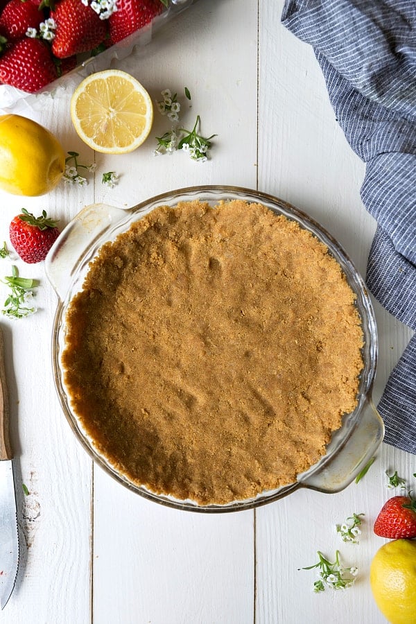 Pie crust in a clear pie pan with fresh lemons and strawberries on the side. 