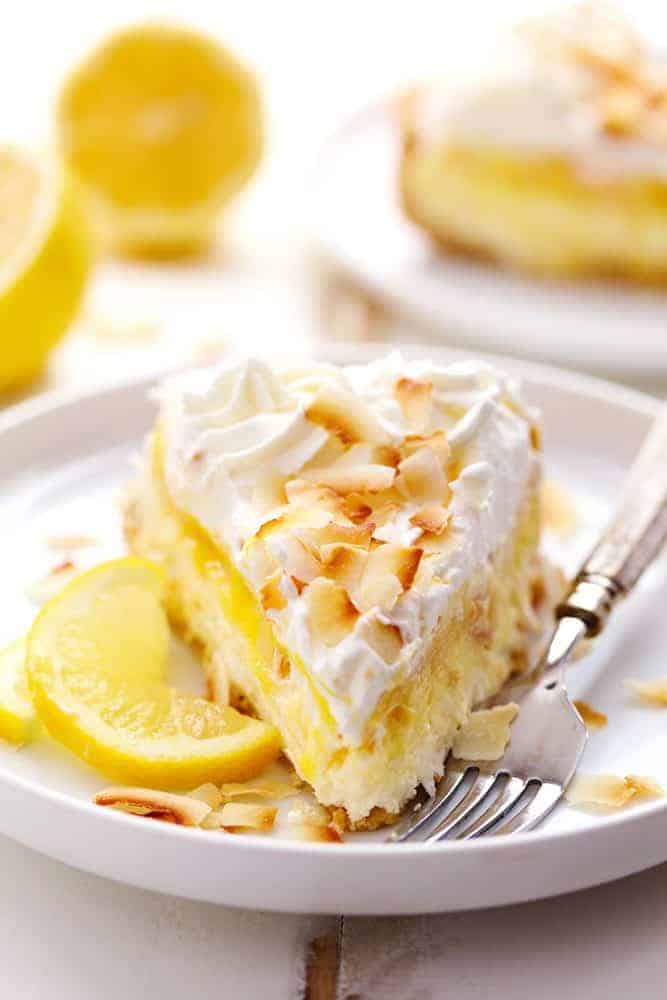 Ein Stück No Bake Lemon Macaroon Cheesecake auf einem weißen Teller mit einer Metallgabel.  Im Hintergrund sind frisch geschnittene Zitronen zu sehen.  