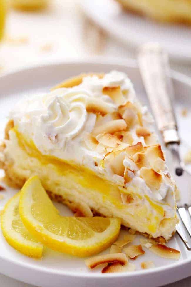 Ein Stück No-Bake-Zitronen-Makronen-Käsekuchen auf einem weißen Teller mit einem Metalllöffel und etwas frisch geschnittener Zitrone als Beilage.  