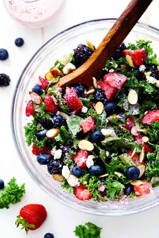 Triple Berry Kale Salad with a Creamy Strawberry Poppyseed Dressing in a clear bowl with a wooden spoon. 