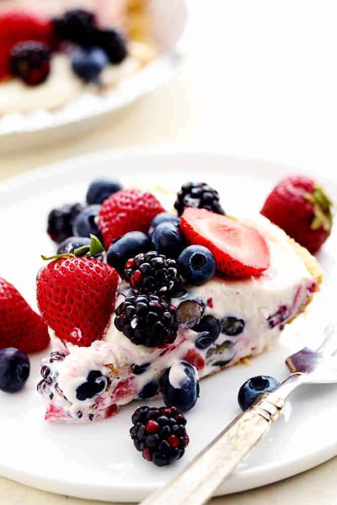 Ein Stück No Bake Very Berry Cheesecake auf einem weißen Teller mit einer Metallgabel und frischen Beeren darauf. 