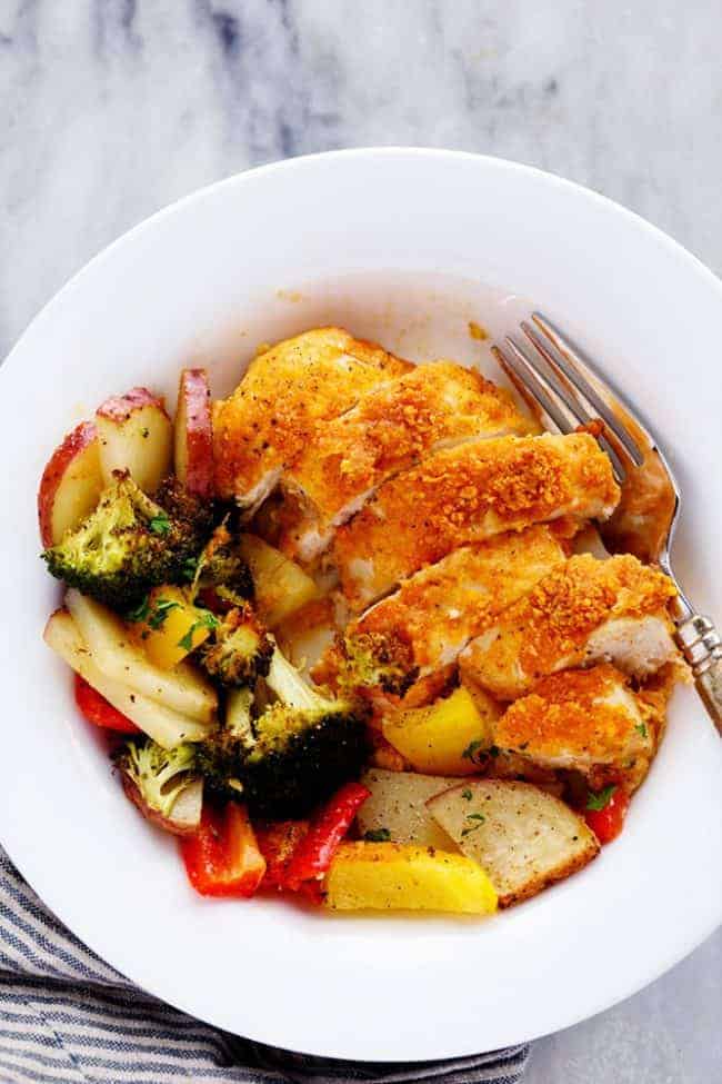 Crispy Parmesan paprika chicken cut into slices and type of vegetables and a white bowl with a fork on the side.