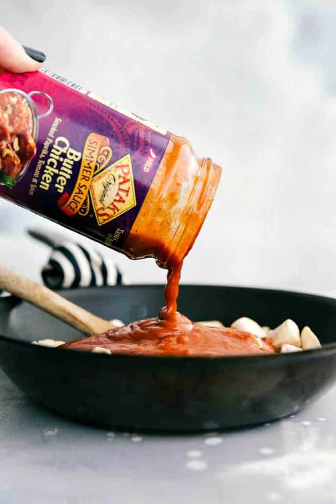 Butter chicken sauce being poured over cooked chicken in a black bowl with a wooden spoon. 